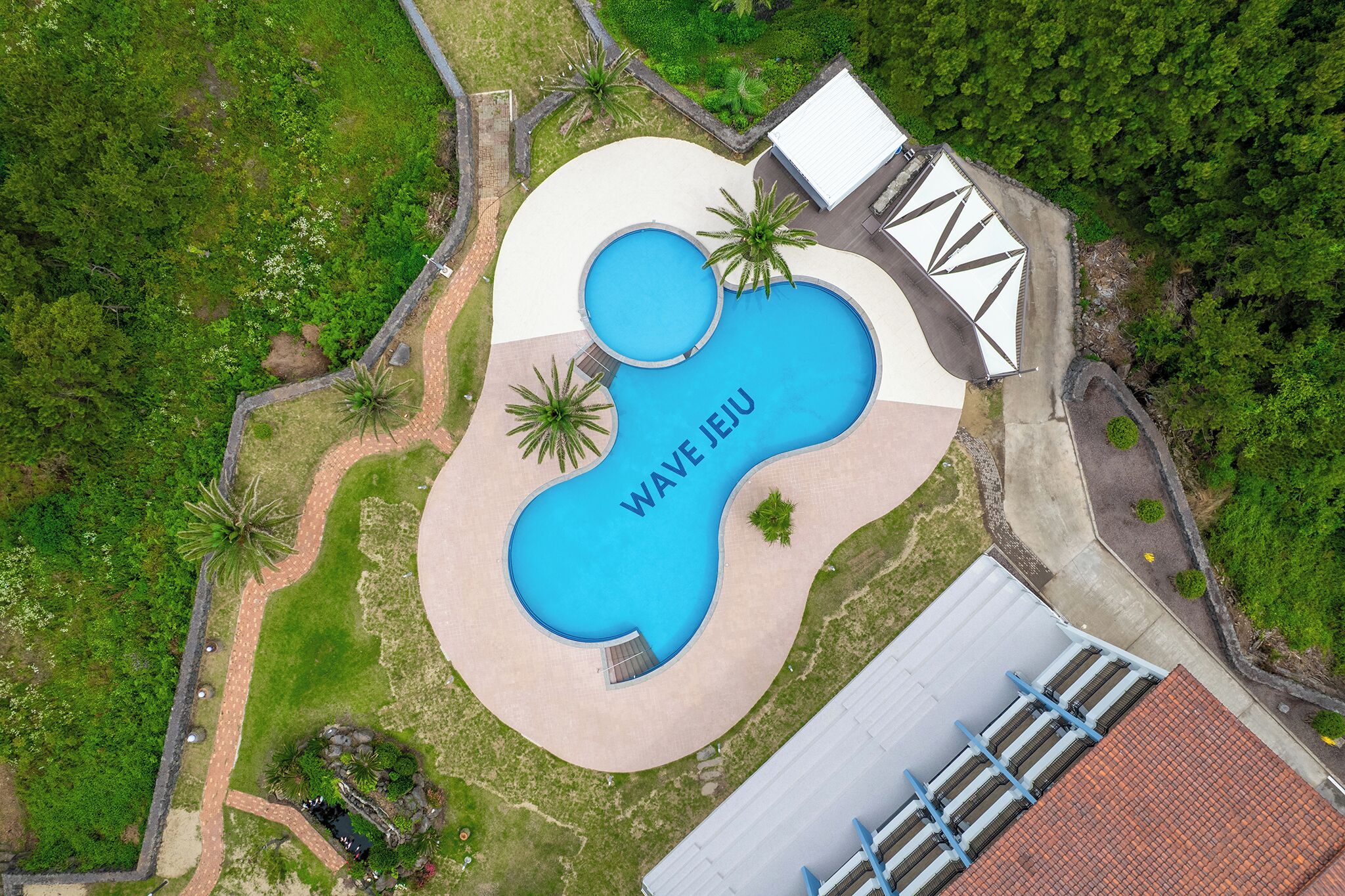 Eine Vogelperspektive zeigt ein Anwesen mit einem großen, wellenförmigen Pool, umgeben von viel Grün und einem Gebäude mit einem Ziegeldach.