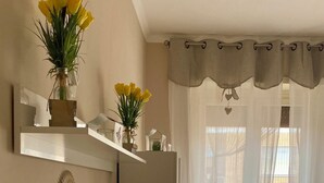 A living room with a flat-screen TV, decorative mirrors, a vase with yellow flowers, and a window with sheer curtains. - Modern and Cozy Oasis in the Heart of the City (Civitavecchia)
