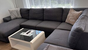 A modern living room with a grey sectional sofa, a glass coffee table, and a view of the outdoors through large windows. - Fantastic vacation apartment (Altendorf)