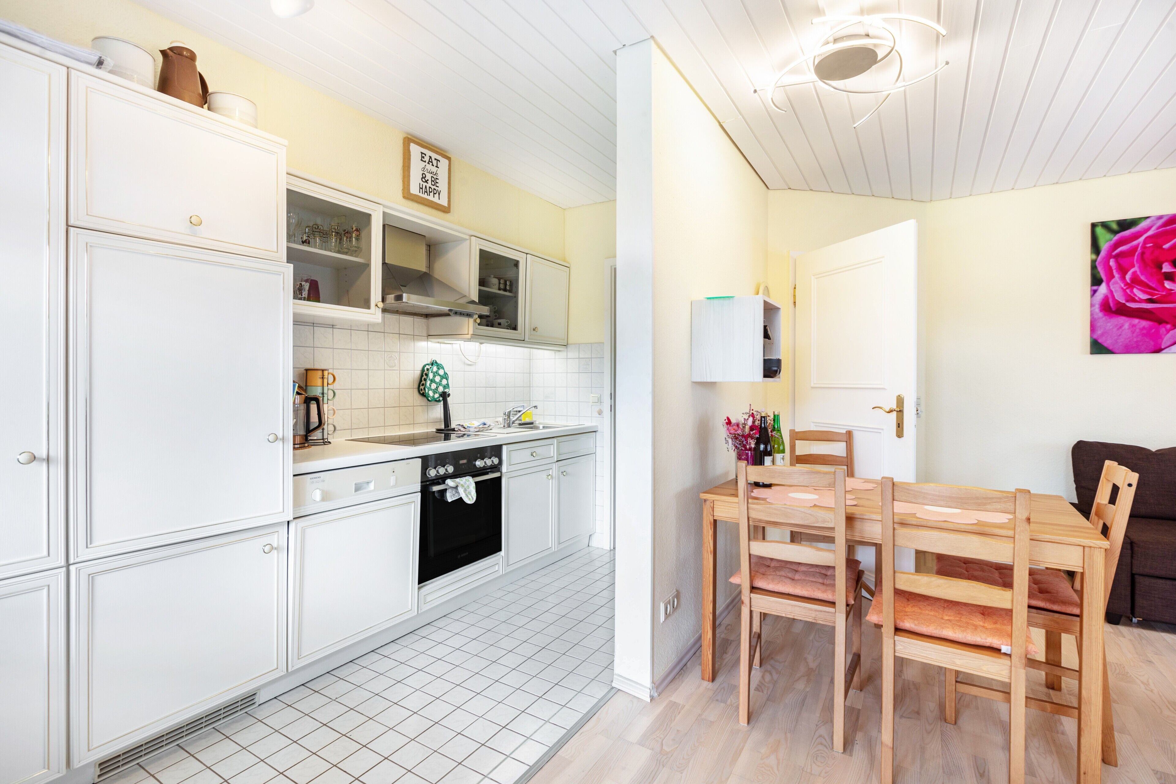 Cocina compacta con muebles blancos, comedor con sillas de madera y una puerta visible que conduce a otra habitación.