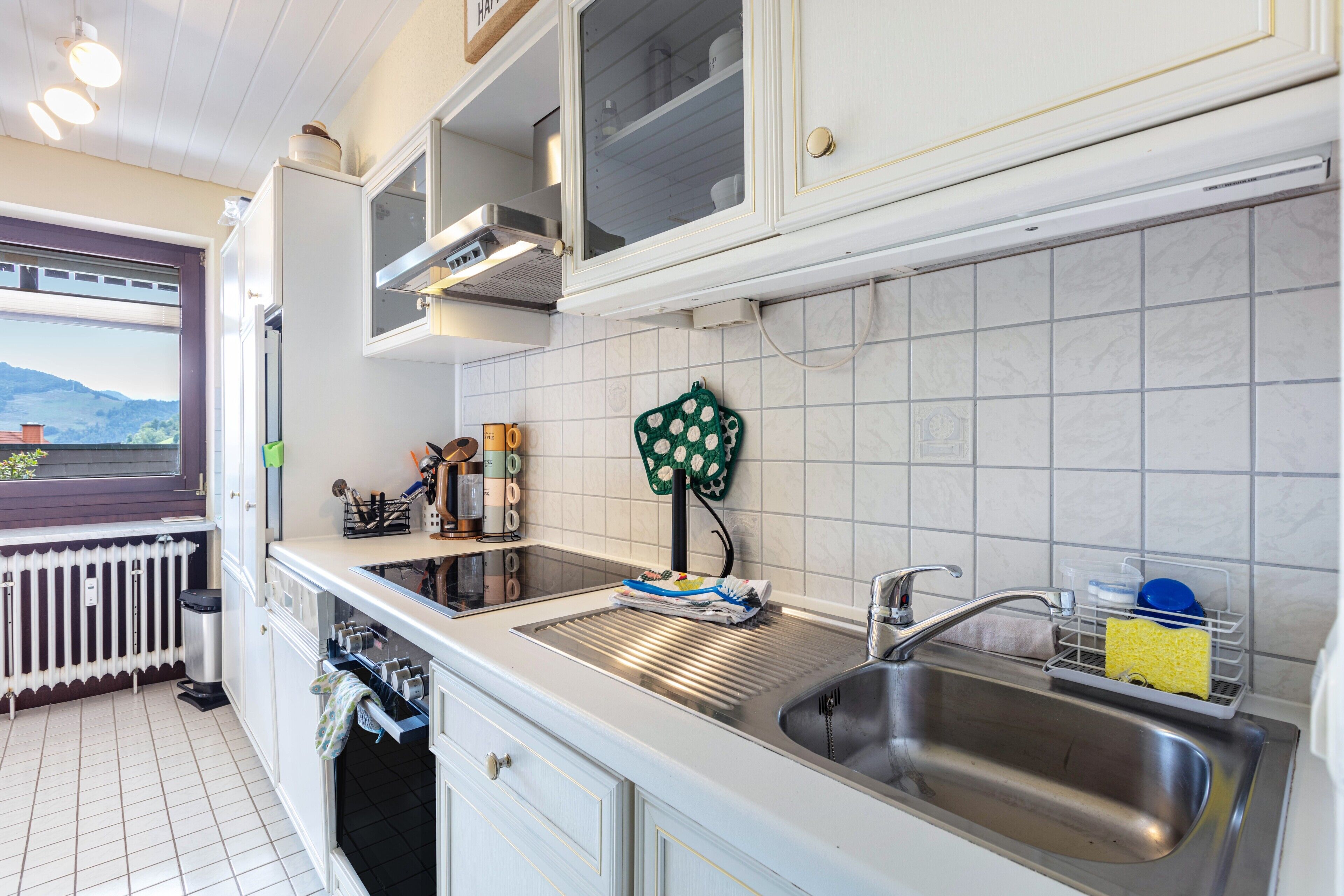 Een keuken met een roestvrijstalen spoelbak, witte kastjes, een raam met uitzicht en een radiator.