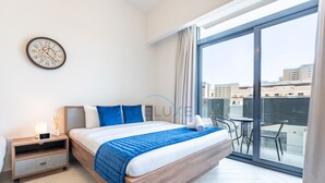 A modern bedroom with a large bed, a bedside table, a clock, and a view of the city through a large window. - Cozy Studio with Peaceful Community Views, Just Steps from Parks & Circle Mall (Dubai)