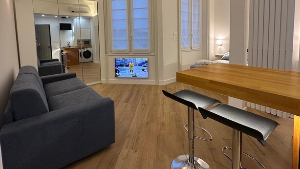 En moderne stue med en grå sofa, et træspisebord og et køkkenområde, der kan ses igennem en glasdør.