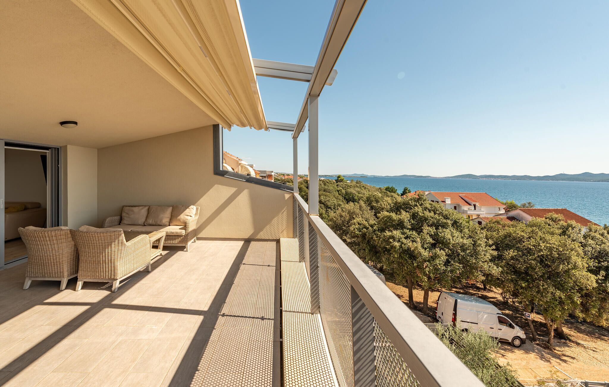 A balcony with wicker furniture, a view of the sea, and a white van parked nearby.