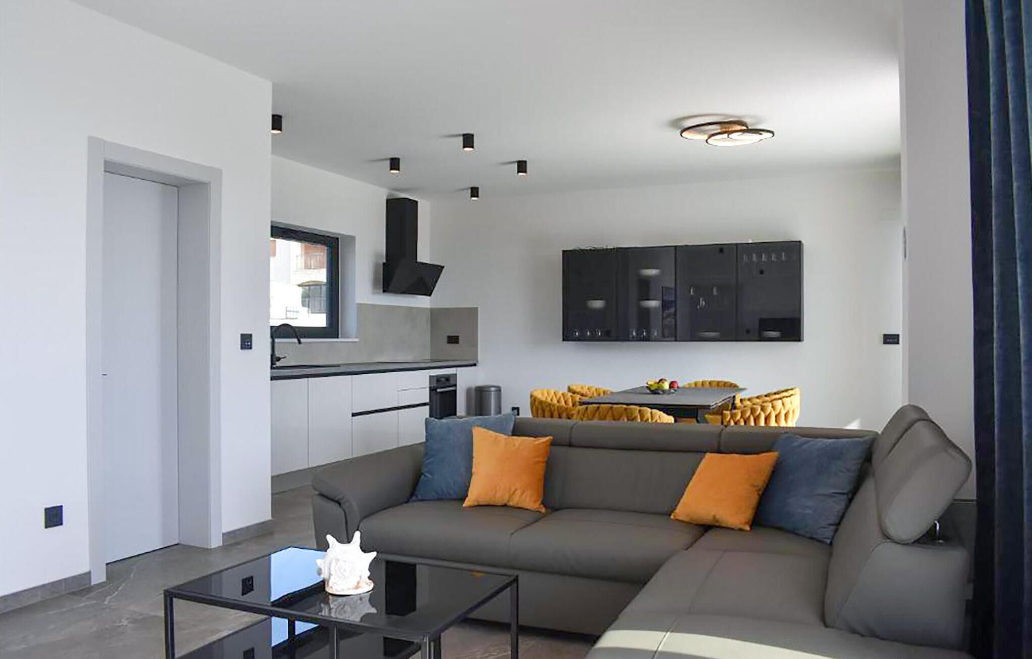 A modern living room with a grey sofa, a glass coffee table, and a kitchen area with white cabinets and a black range hood.