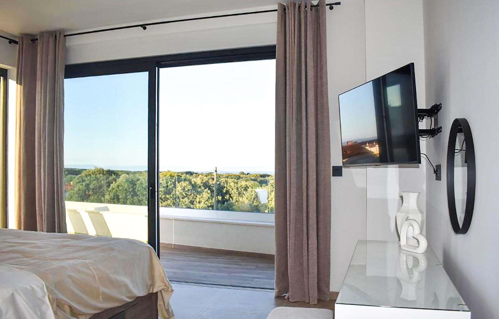 Et hotelværelse med seng, fjernsyn og balkon med udsigt til træer og bygninger.