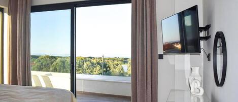 Et hotelværelse med seng, fjernsyn og balkon med udsigt til træer og bygninger.