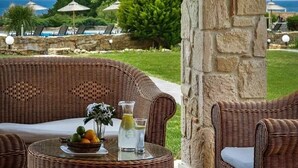 A covered outdoor seating area with wicker furniture, a table with a fruit bowl and drinks, and a stone pillar. - Evlogia Village by Greek Pride (Nea Skioni)