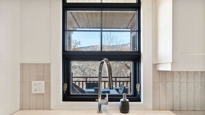 A modern kitchen with a stainless steel sink, a black faucet, and a window overlooking a deck. - Les Eaux: Luxe Mountain View Condo + Private Sauna (Mont-Tremblant)