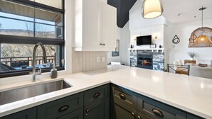 A modern kitchen with a large window, a stone fireplace, and a dining area with a TV above it. - Les Eaux: Luxe Mountain View Condo + Private Sauna (Mont-Tremblant)