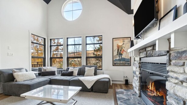 A modern living room with a stone fireplace, large windows, and a sectional sofa. - Les Eaux: Luxe Mountain View Condo + Private Sauna (Mont-Tremblant)