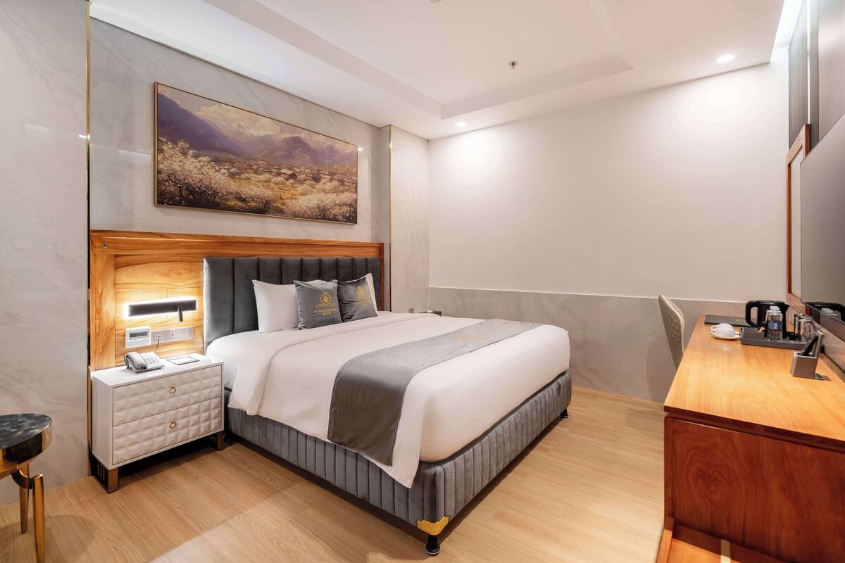 Ein modernes Hotelzimmer mit einem großen Bett, einem Holzkopfteil, einem Nachttisch mit Lampe, einem Schreibtisch mit Kaffeemaschine und einem großen Landschaftsbild an der Wand.