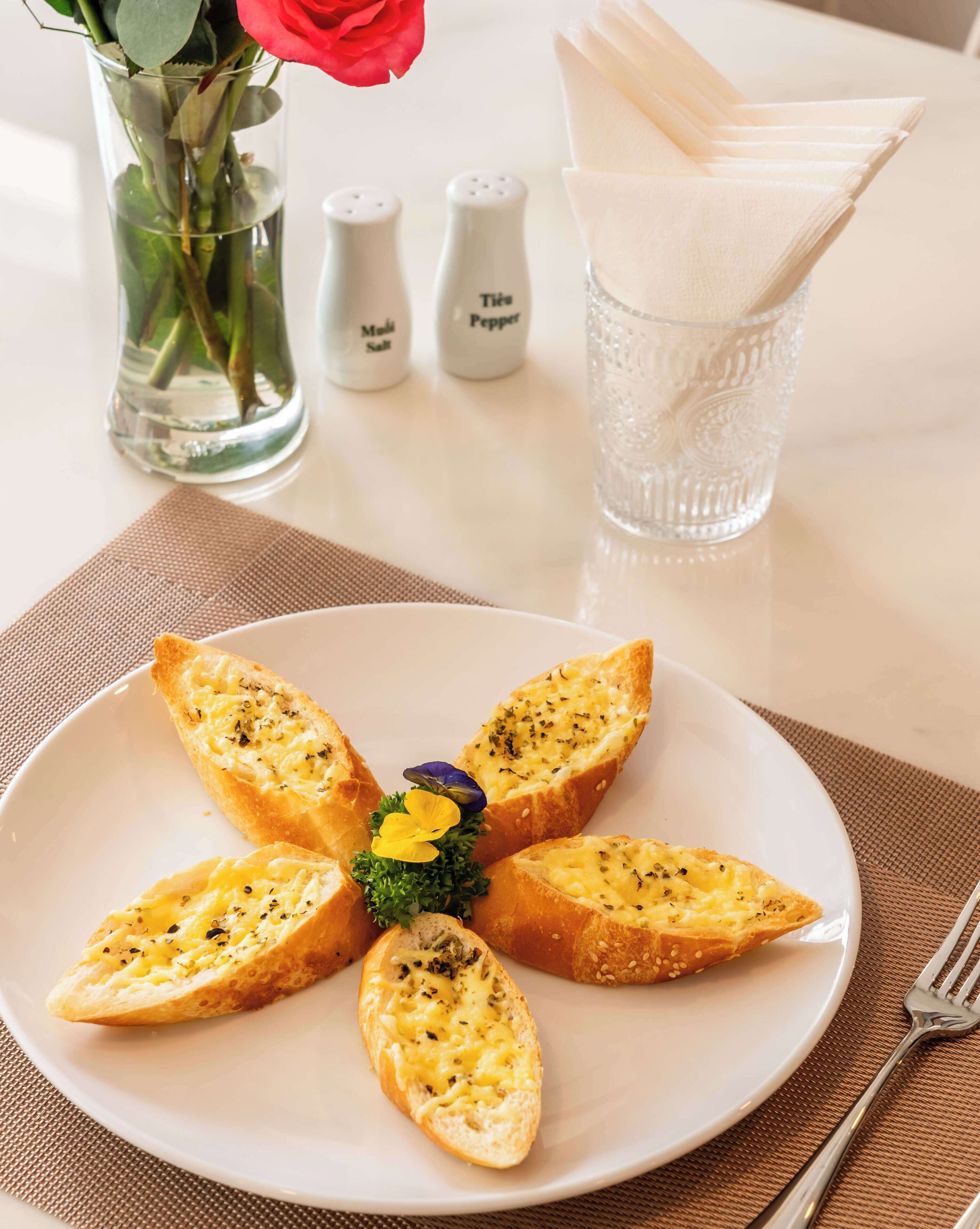 Um prato de pão de alho decorado com ervas e uma flor, saleiro e pimenteiro, um copo de água e uma rosa em um vaso sobre uma mesa.