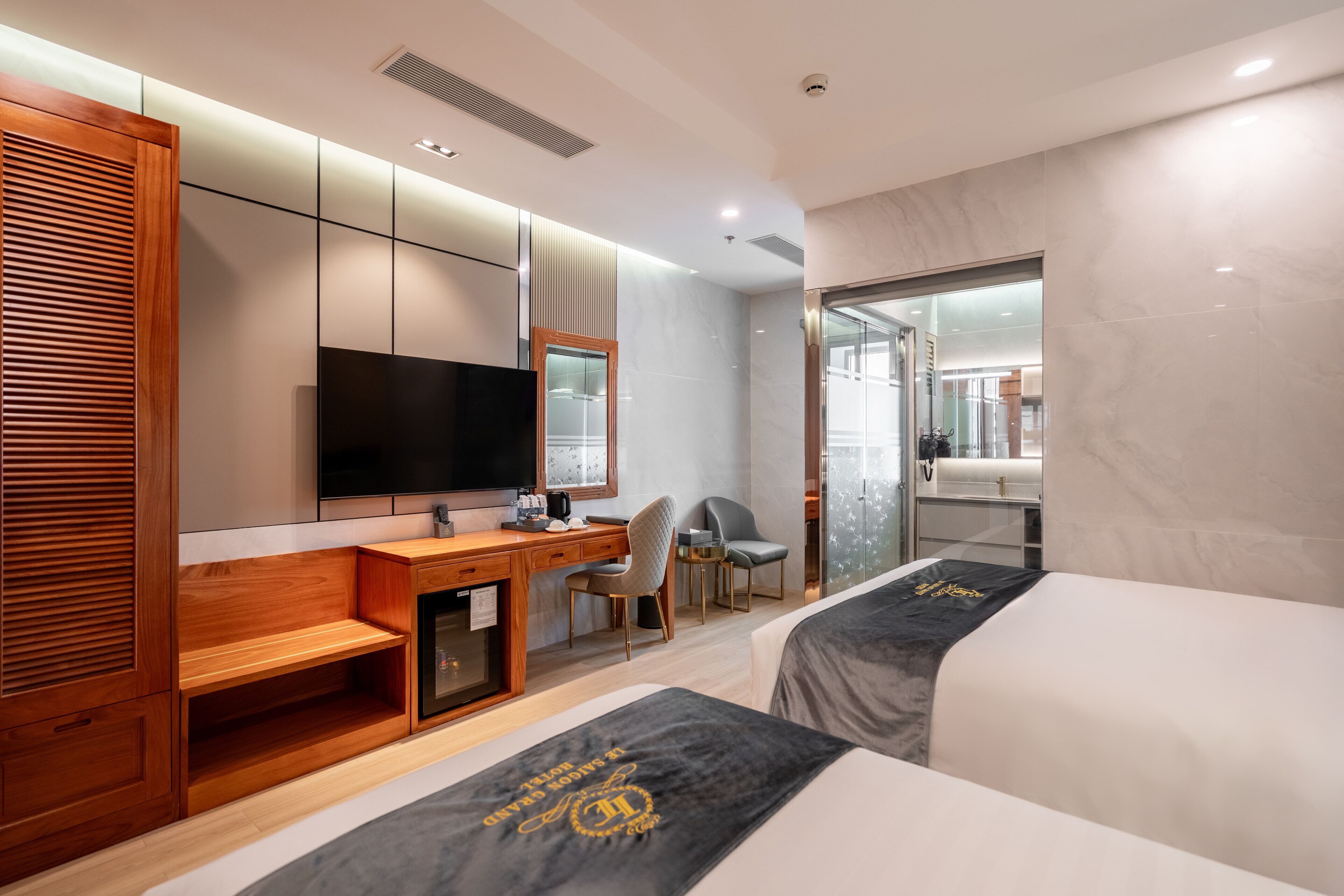 A modern hotel room with a wooden desk, a flat-screen TV, a bed with a black and gold bedspread, a chair, and a large mirror.