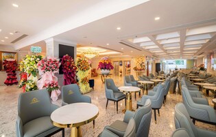 A hotel lobby with a modern interior, featuring a chandelier, multiple seating areas with round tables, and a variety of floral arrangements.