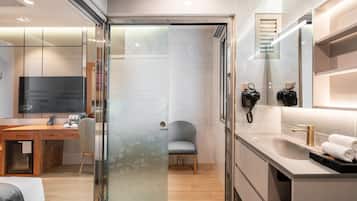 A modern hotel room with a glass-enclosed shower, a wooden desk, a chair, a large mirror, and a vanity with a gold faucet.