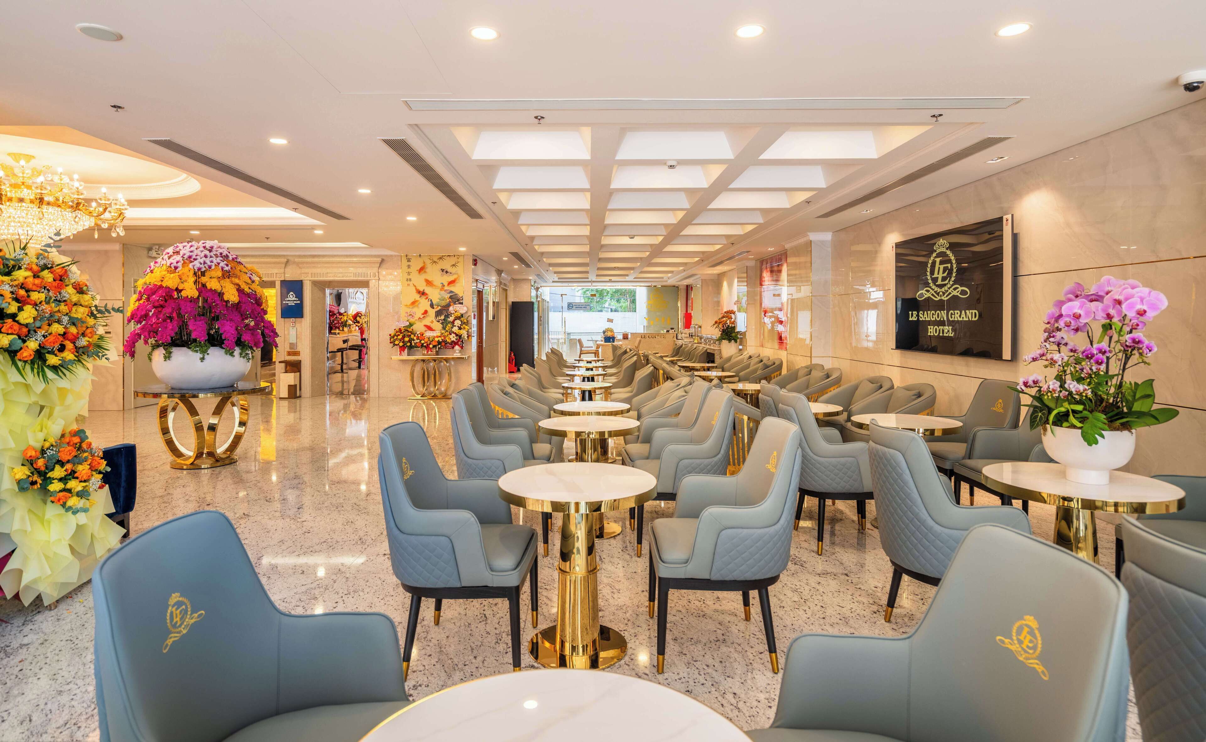 A hotel lobby with a long row of tables and chairs, a large floral arrangement, and a sign for Le Saigon Grand Hotel.