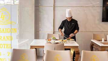 A hotel dining area with a chef preparing food. There are multiple dining tables with chairs, each with a logo on the backrest. The walls are adorned with a marble-like pattern.