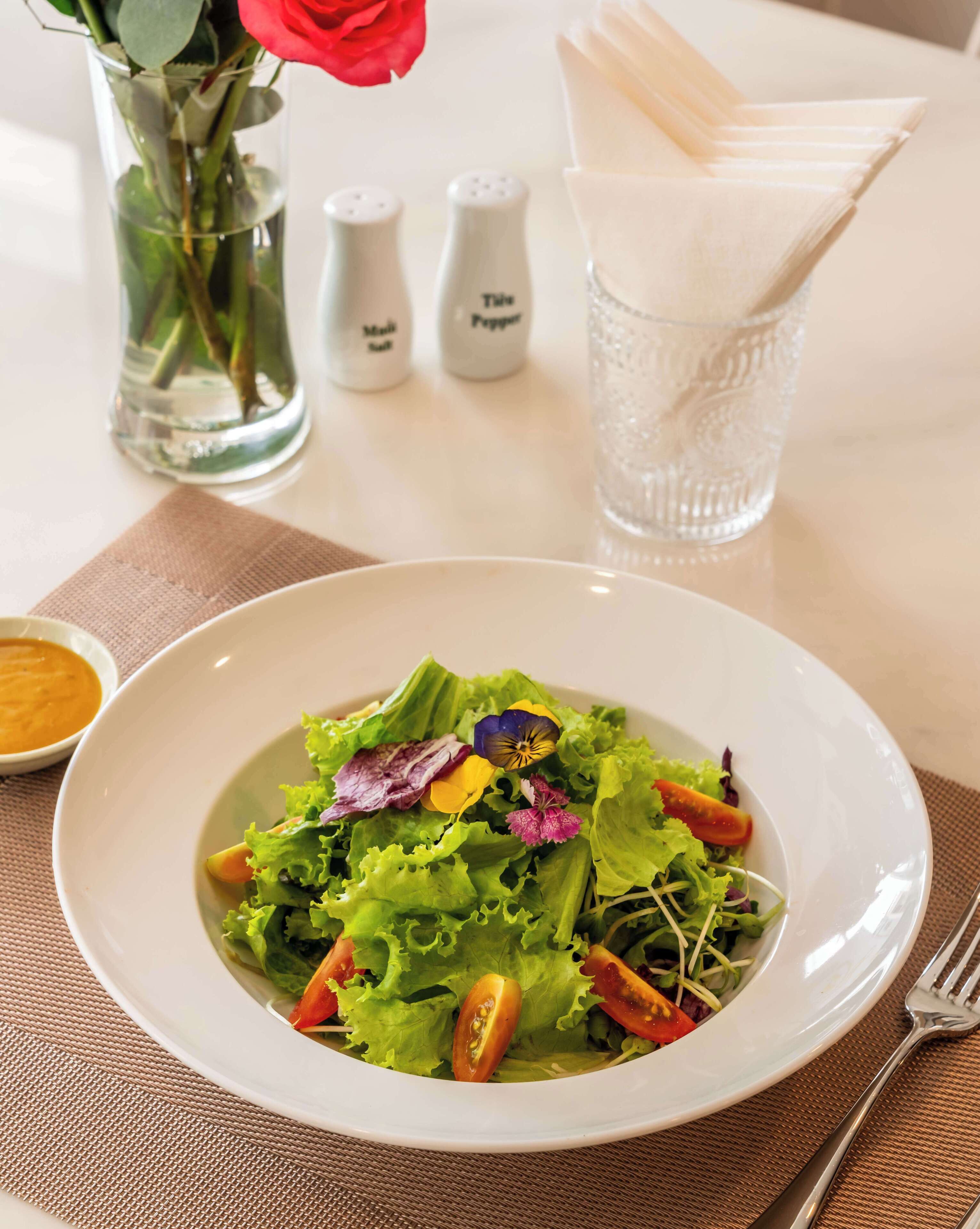 A white plate with a salad, a glass of water, salt and pepper shakers, a vase with a rose, and a napkin holder on a table.