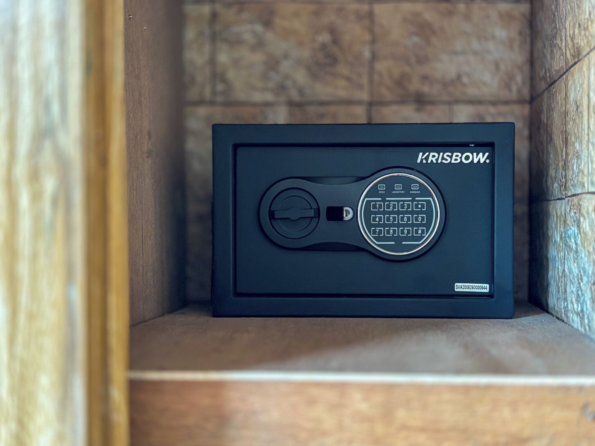 A black safe with a keypad and a lock mechanism, branded 'KRISBOW,' placed in a corner with a tiled wall background.