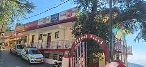 A hotel entrance with a gated driveway, a signboard advertising 'Hotel Deodar Villa' and 'make my trip' services, and parked cars. - Deodar Villa (Dharamshala)