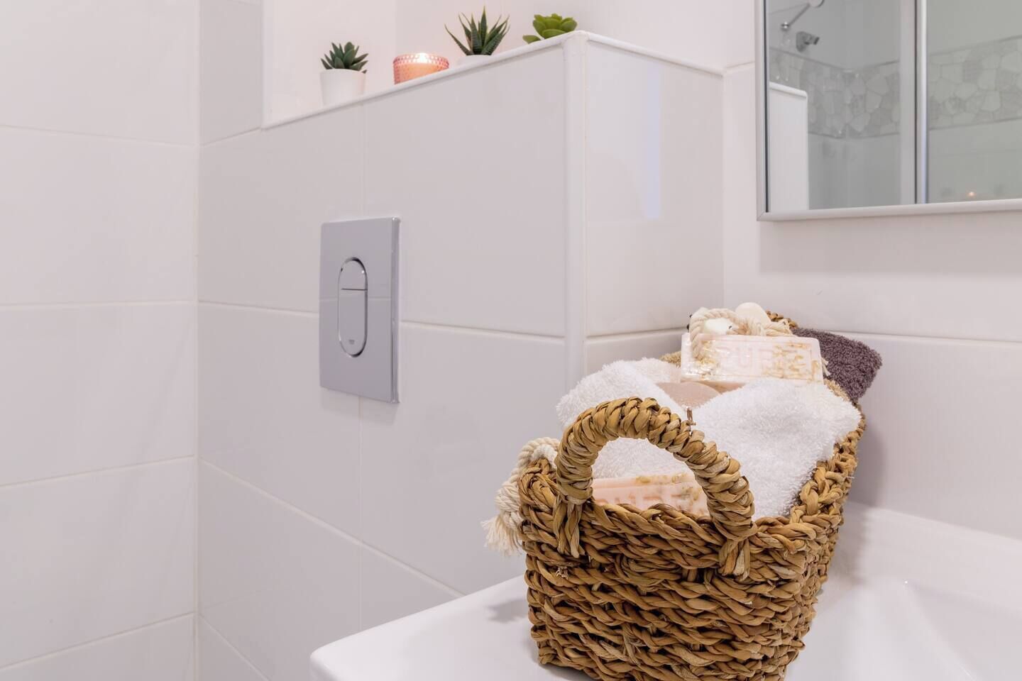 Een badkamer met witte tegels, een zilveren lichtschakelaar, een rieten mand met handdoeken en een klein plankje met planten en een kaars.