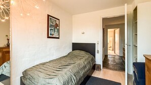 Un dormitorio con una cama individual, un escritorio, una silla, una puerta que da a otra habitación y una lámpara fijada a la pared.