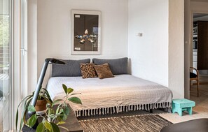A bedroom with a bed, a nightstand, a lamp, a plant, a mirror, and a view of the outside.