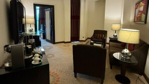 Ein Hotelzimmer mit Flachbildfernseher, einer Kaffeemaschine, einem Schreibtisch, einer Couch, einem Sessel und einer Lampe.