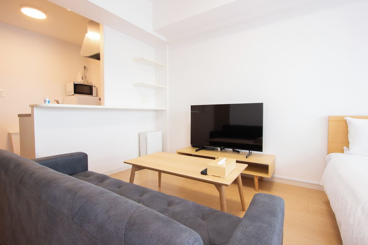 Ein modernes, minimalistisches Wohnzimmer mit grauem Sofa, einem Holztisch, einem Flachbildfernseher und einem kleinen Beistelltisch.