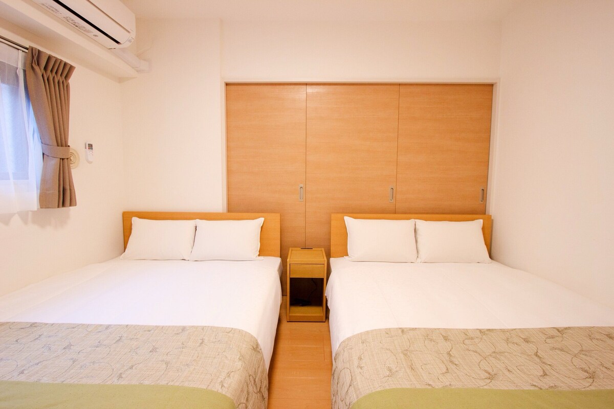 A hotel room with two single beds, a wooden wardrobe, a bedside table, and an air conditioning unit.