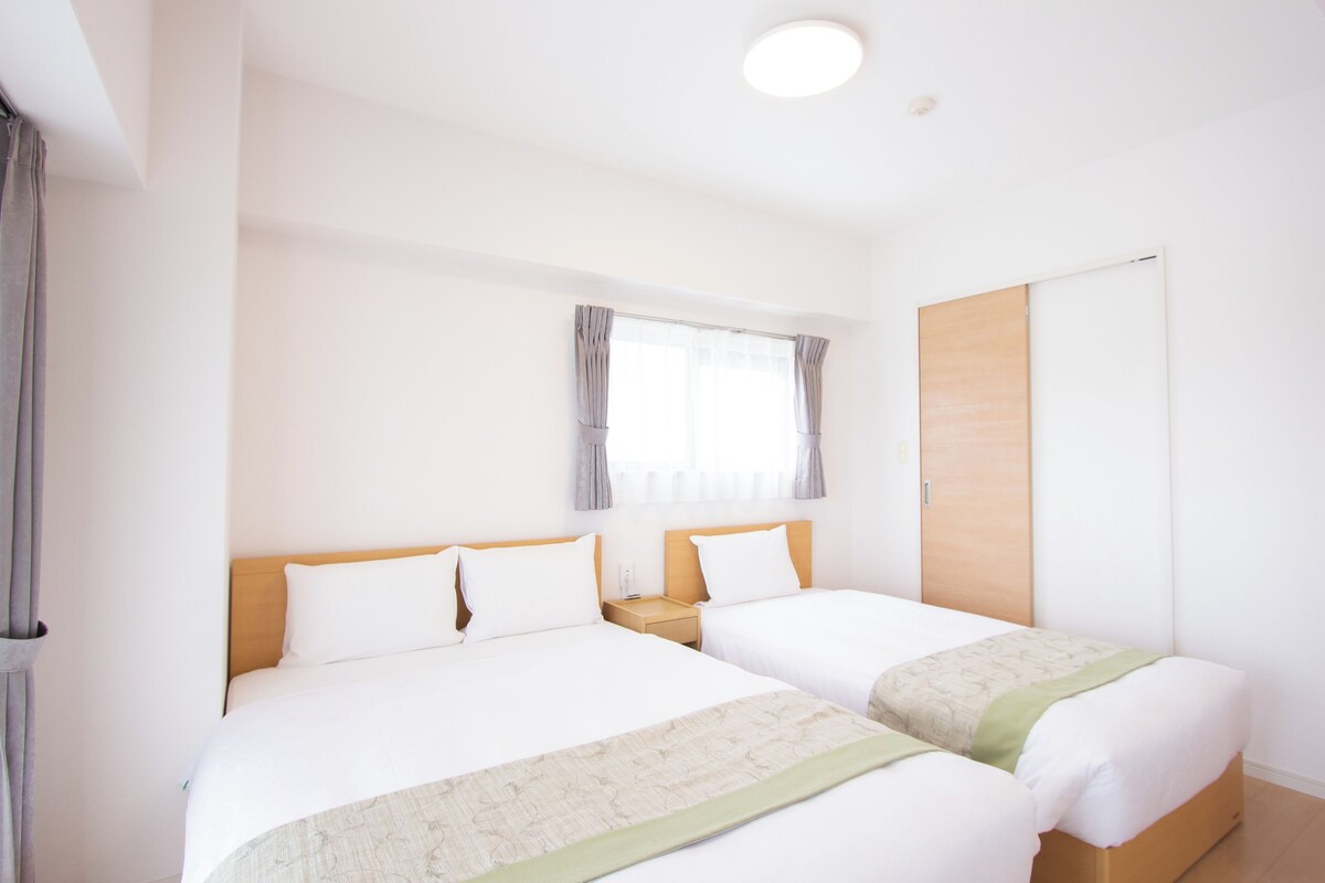 A hotel room with two beds, a window with curtains, a wooden wardrobe, and a nightstand with a lamp.