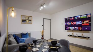 Una sala de estar moderna con comedor, un televisor de pantalla plana mostrando varios canales, un sofá con cojines y un jarrón decorativo.