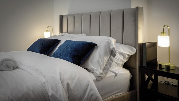 Una cama bien hecha con almohadas blancas y azules, un cabecero tapizado en gris y lámparas de noche sobre una mesita.
