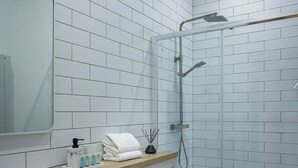 Un baño moderno con paredes de azulejos blancos, un box de ducha de vidrio, un inodoro blanco y un tocador con espejo y artículos de aseo.