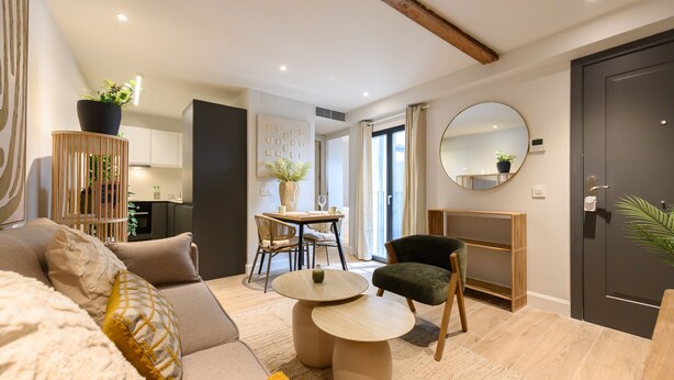 A modern living room with a sofa, a round table, and a chair. There is a dining area with a table and chairs in the background.
