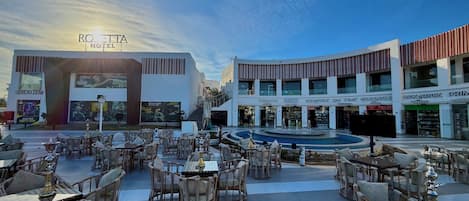 An outdoor seating area with tables and chairs, a pool, and a hotel entrance with signage.