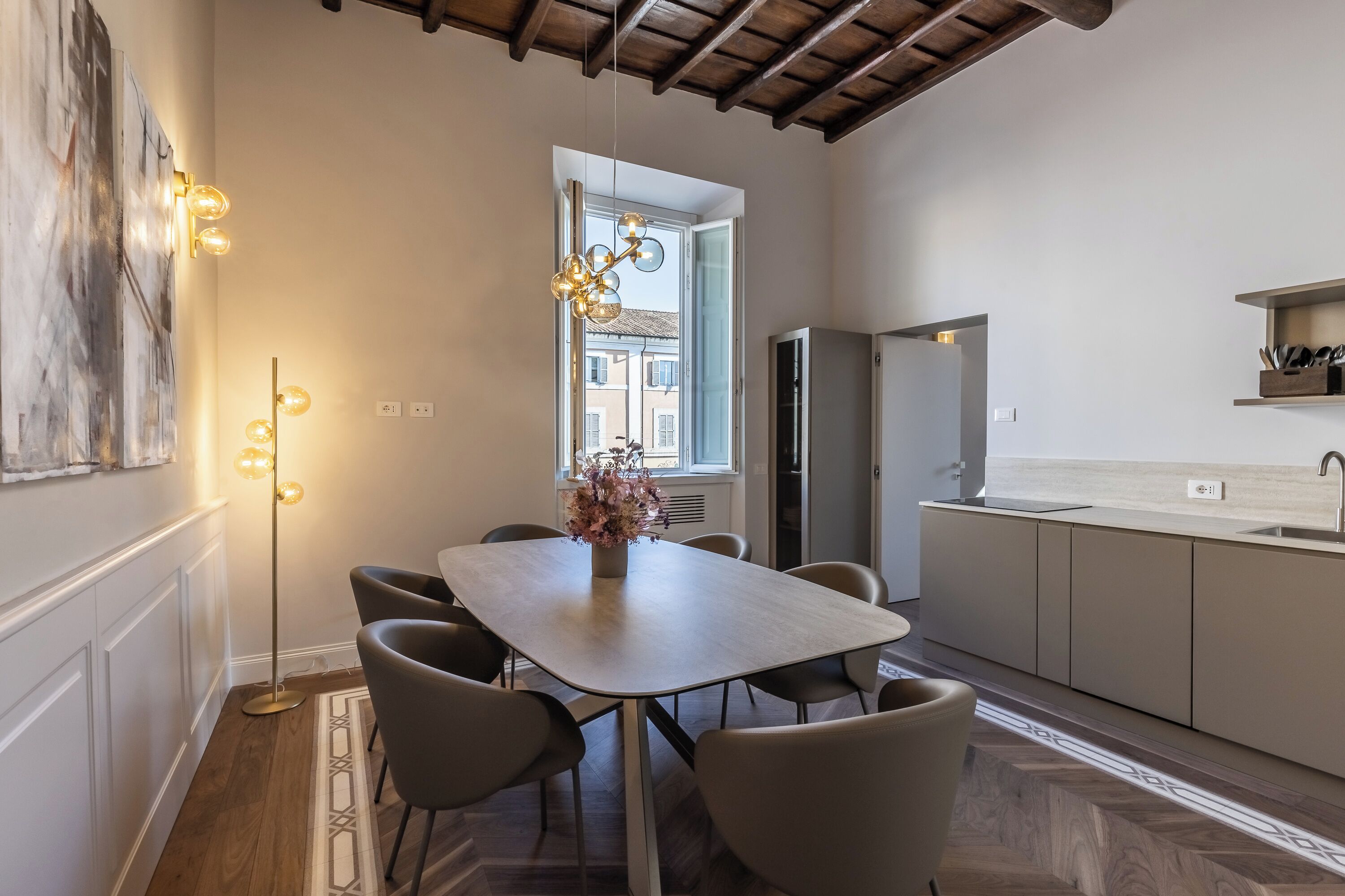 A modern kitchen with a dining area, featuring a wooden dining table, chairs, a pendant light, and a view through a window.
