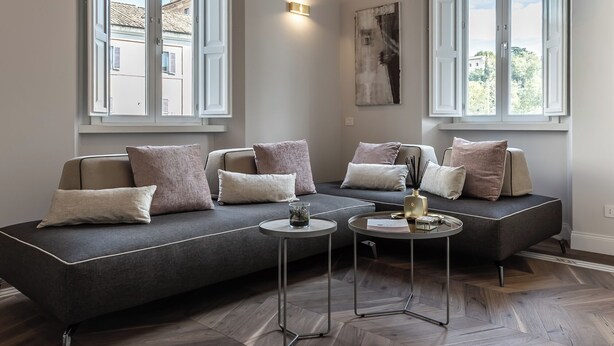 Una sala de estar moderna con un sofá seccional grande, dos ventanas con contraventanas blancas, piso de madera en patrón de espiga y una lámpara colgante.