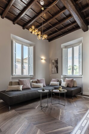 A modern living room with a large sectional sofa, two windows with white shutters, a wooden herringbone floor, and a hanging light fixture. - Porta Portese Palace (Rome)