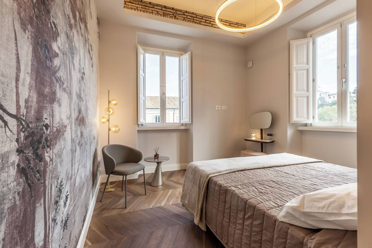 A modern bedroom with a large bed, a chair, a small table, and a wall-mounted mirror.