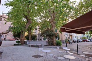An outdoor café with tables and chairs, surrounded by trees and a building with a staircase.