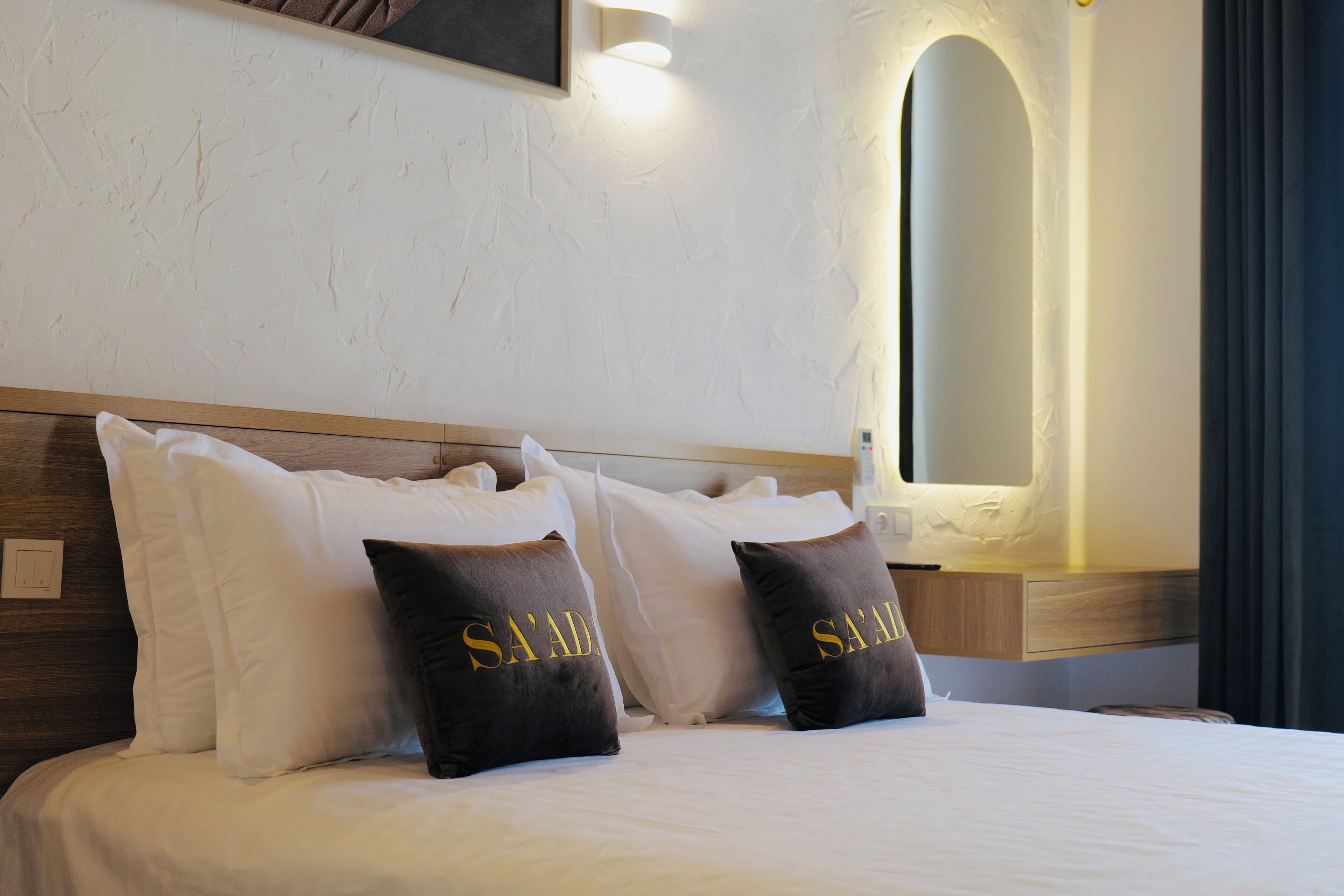 Habitación de hotel con cama, almohadas blancas y marrones, cabecero de madera, lámpara de pared y espejo.