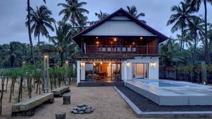 A two-story house with a balcony, a swimming pool, and a sandy area with benches and rocks.