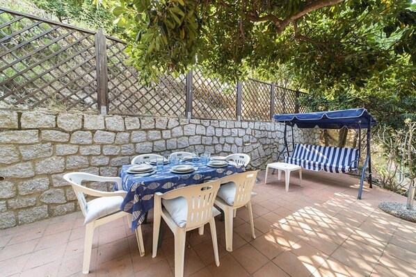 Een terras buiten met een stenen muur, een eettafel en een blauw gestreepte luifel.