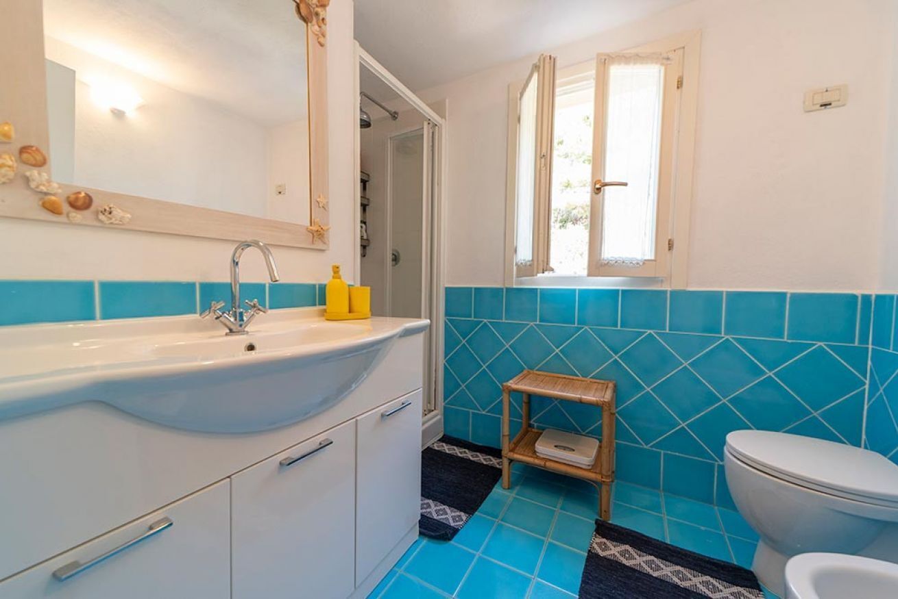 Un baño con un lavamanos blanco, azulejos azules, un estante de madera y una ventana.