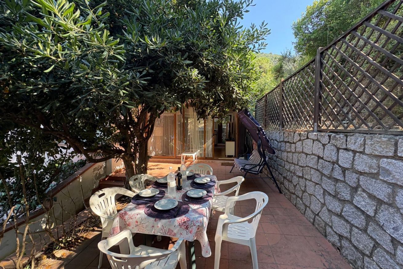 An outdoor dining area with a table set for four, surrounded by white chairs, under a tree.