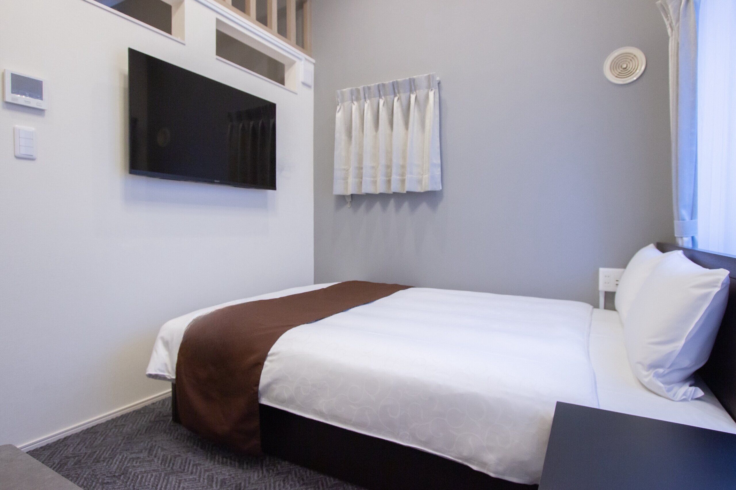 A hotel room with a bed, a flat-screen TV mounted on the wall, a window with curtains, and a thermostat on the wall.
