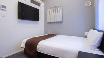 A hotel room with a bed, a flat-screen TV mounted on the wall, a window with curtains, and a thermostat on the wall.