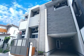 A modern multi-story building with a covered entrance, air conditioning units, and a gated area. - HOTEL FAMILIA Hakata East (Fukuoka)